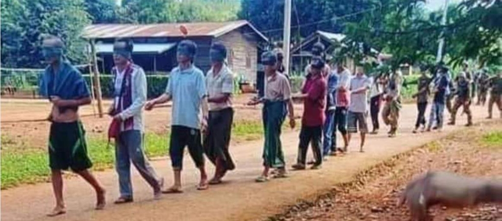 눈을 가린 채 줄에 앞뒤로 묶여 걸어가는 미얀마 시민들. 뒤로 군인들이 보인다.